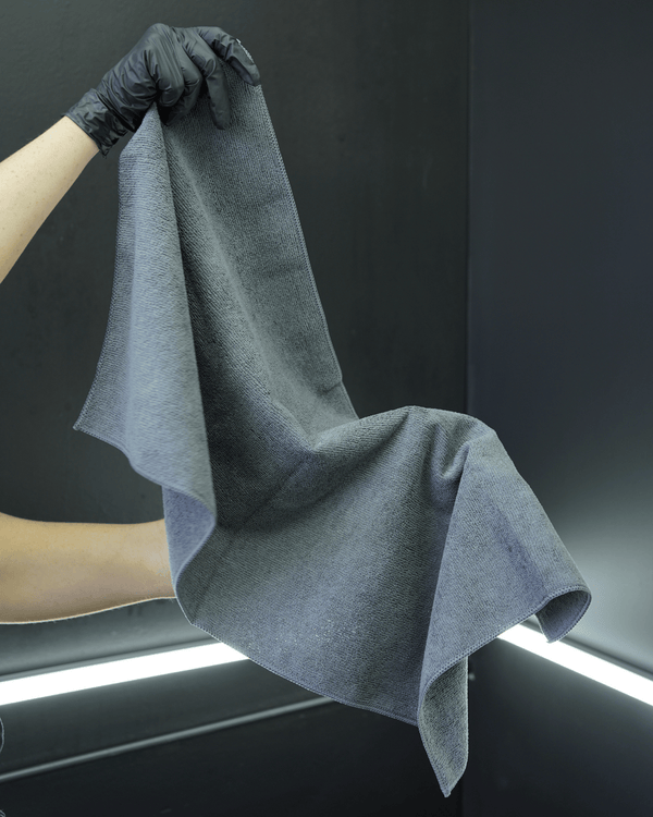 A person wearing black gloves holding a large grey microfibre towel on a dark background