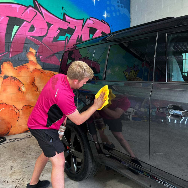 a person in a pink shirt using a yellow microfibre cloth to detail a car