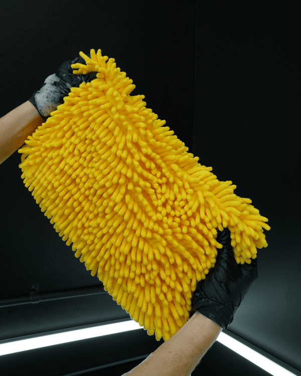 A person wearing black gloves holding a large yellow noodle sponge on a dark background