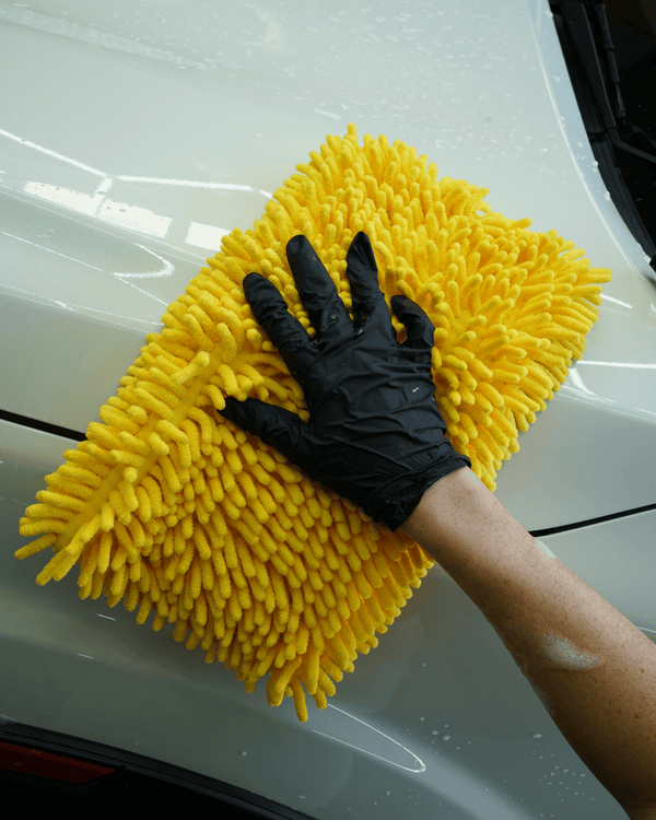 A person wearing black gloves using a large yellow noodle sponge to clean a white car