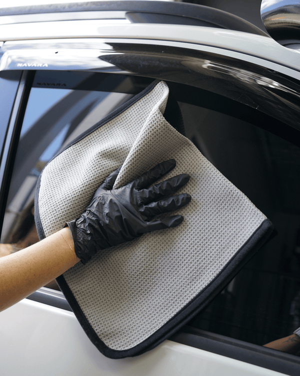 A person wearing black gloves cleaning a car window with a grey waffle weave towel with a back border.