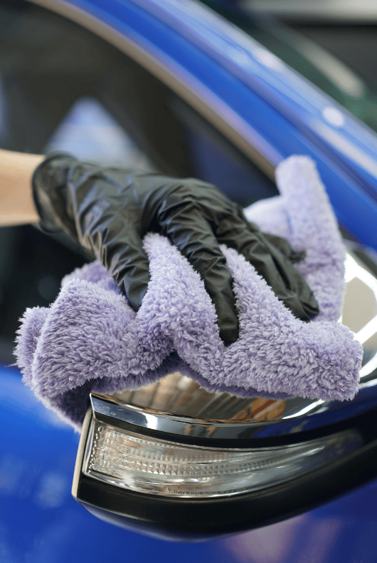 A person cleaning a blue car mirror with a purple microfibre cloth and black gloves on