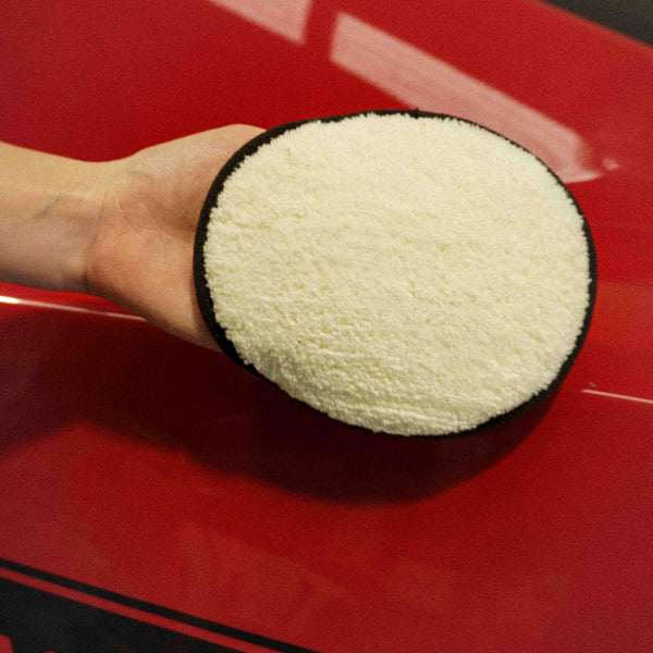 A person holding a microfibre applicator pad over a red car bonnet