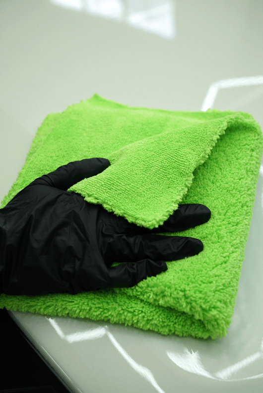 A person cleaning a white car with a green microfibre cloth and black glove on