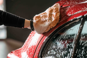 A person using a soapy orange microfibre mitt to wash a red car