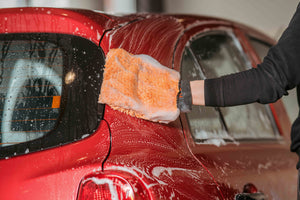 A person using a soapy orange microfibre wash mitt to wash a red car
