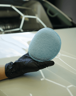 A person wearing a black glove holding a round applicator pad on a white car