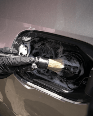 A person holding a detailing brush cleaning a car fuel cap with black gloves on