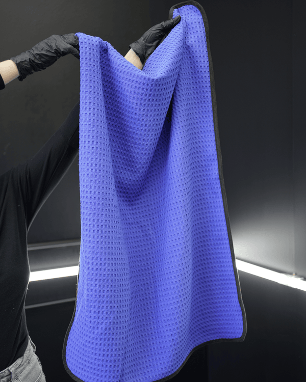 A person wearing black gloves holding a large blue waffle weave car drying towel in a detailing bay
