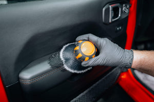 Hand in grey glove using ADBL Fat Brush to clean car door panel armrest, agitating foam on black vinyl/leather
