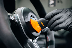 ADBL detailing brush with soft orange bristles cleaning a BMW steering wheel emblem and dash buttons