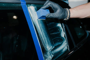 Close-up of a hand polishing car glass with ADBL Glass Polish and a blue applicator, taped area visible