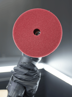 Red polishing pad held by a gloved hand against a neutral background