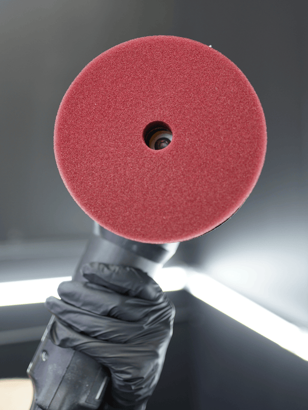 Red polishing pad held by a gloved hand against a neutral background