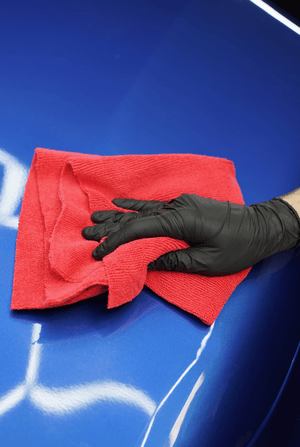A person cleaning a blue car with a red microfibre cloth and black gloves on.