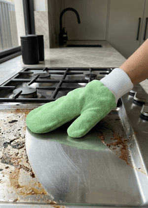 A green all-purpose microfibre cleaning mitt being used to clean a dirty stainless steel stove top