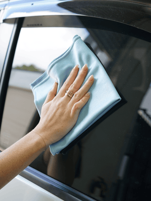 Hand cleaning a car window with a blue microfibre cloth.