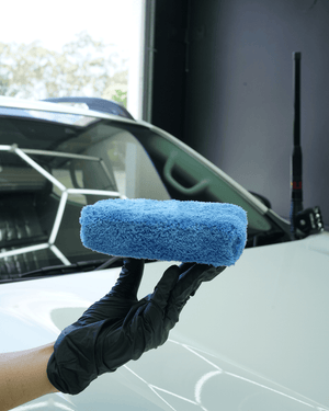 A person wearing black gloves holding a plush blue microfibre applicator next to a white car