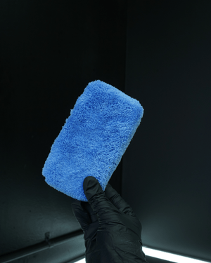 A person wearing black gloves holding a blue plush microfibre applicator on a dark background