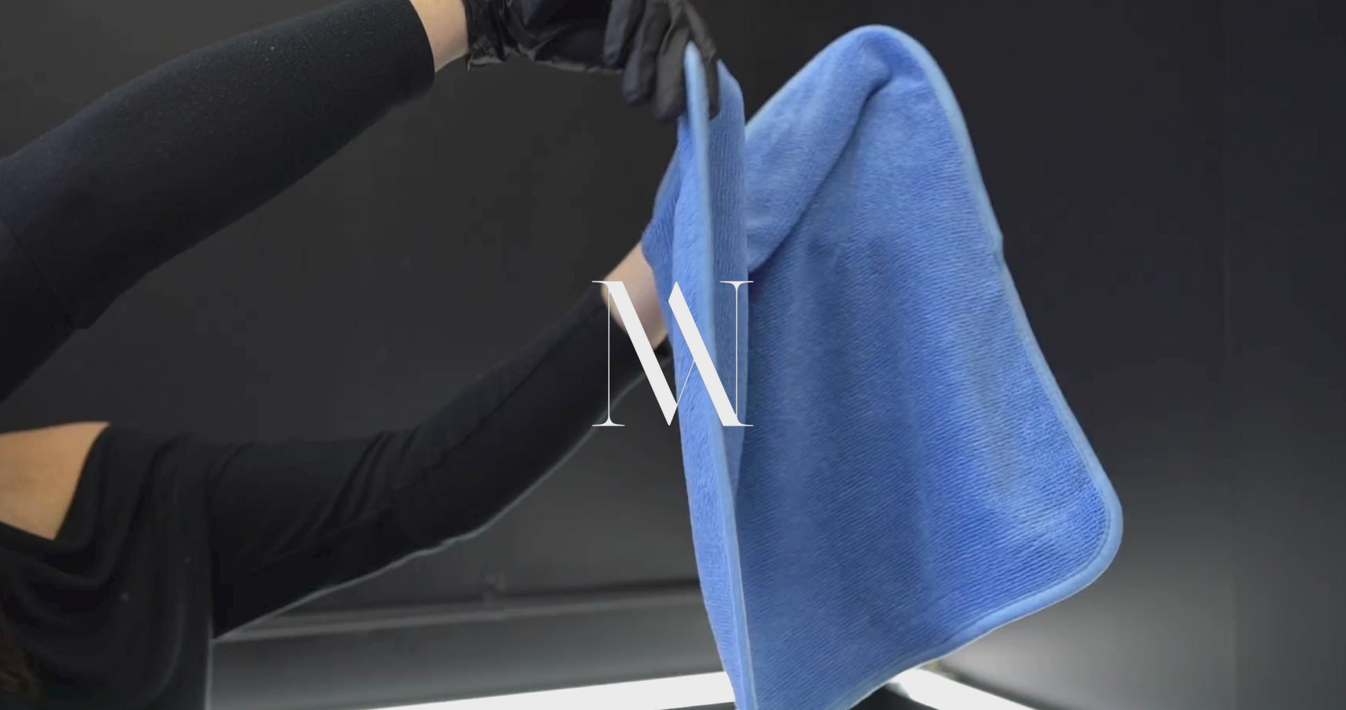 A person wearing black gloves holding a blue microfibre drying towel in a detailing bay